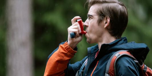 Young man on hike holding inhaler to his mouth with right hand