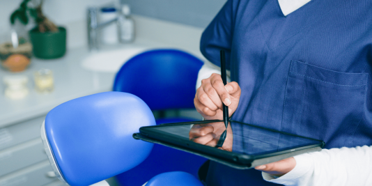 A dental hygienist holds a tablet in the operatory while review the screen and notating