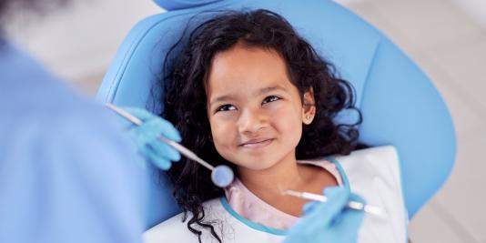 Child wtih long dark hair sits in dental chair smiling at hygienist who is holding dental intruments in her gloved hands.