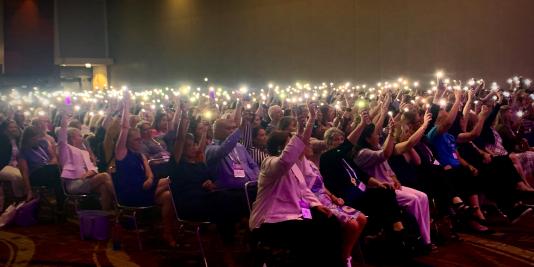 Darkened ballroom with seated audience holding phone lights up in the air
