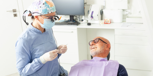 Hygienist with scrub cap, mask and loupes speaking with older gentleman in glasses with lilac paper bib in dental operatory chair