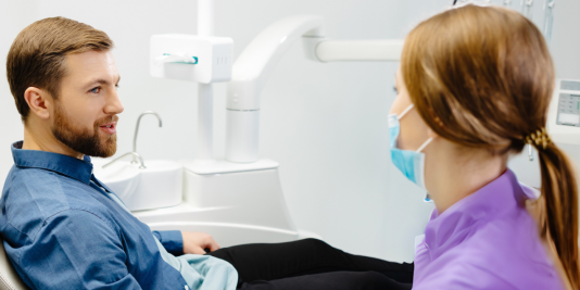 Hygienist in purple shirt facing and speaking with bearded man in dental operatory chair