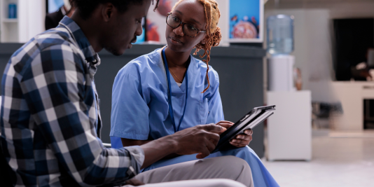 Young female hygienists in blue scrubs holding tablet seated and speaking with male patient in waiting area.